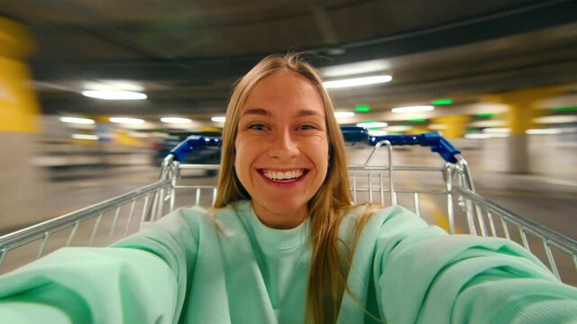 Excited happy young woman laugh and smile, sit inside shopping cart at parking lot of shopping mall or center. Generation z millennial teenager have fun on weekend, record selfie for social media