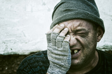 Portrait of a homeless man in a knitted hat, who is crying with his hand to his face.