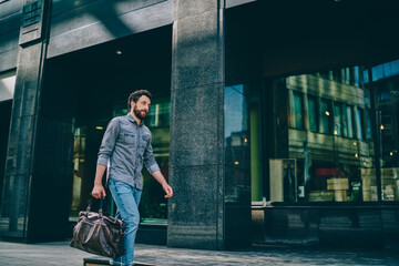 Bearded hipster guy dressed in casual wear holding bag in hand strolling on street in urban setting near big mall.Stylish young man 20 years old walking outdoors and going to sport gym