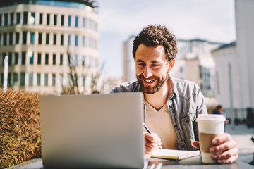 Cheerful bearded young man laughing while watching tutorial online on website on modern laptop computer using wireless internet and writing down notes sitting outdoors in cafe with tasty coffee to go