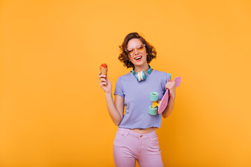 Spectacular white woman with short hair posing with ice cream. Refined european girl with skateboard isolated on orange background.