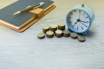 Stack of coins, alarm clock, and notebook on wooden table 