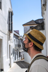 Photo of a young and attractive guy wearing a hat and a reusable face mask travelling and visiting a city. Tourism during coronavirus outbreak. Alone in the city