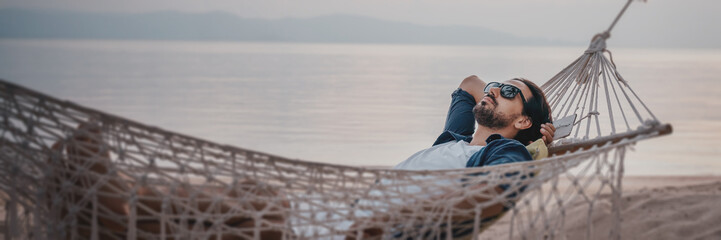 Young handsome Latin man in sunglasses relaxing in a hammock on the beach at sunset on the beach, banner format