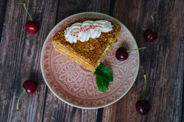 Piece of multi-layer cake with cream on a plate. Classic honey cake pieces with custard. Homemade honey cake on wooden background with cherry and mint leaves. Delicious slice of layered tart on plate