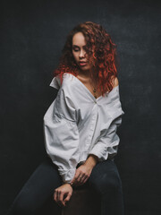 Portrait of a young woman with red and curly hair in a white shirt on a dark background. A girl in jeans is sitting on a chair and posing in a photo Studio.Look down, photo in a dark key