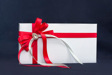 The gift box is white with a beautiful red bow. Gift on a dark background. Holidays and surprises. Satin bows with rhinestones.
