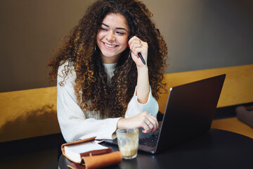 Cheerful attractive young blogger uploading multimedia files on web site enjoying working process in coffee shop, smiling skilled freelancer having fun doing remote job using modern technology