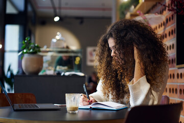 Pensive female student with curly hair concentrated on learning process writing report in cafe...