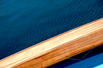 Varnished wooden side deck of a sailing boat, visible small section of light blue cockpit and large area of dark blue water overboard.