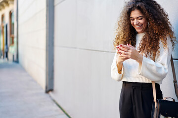 Attractive cheerful casually dressed hipster girl having fun outdoors watching funny video via smartphone, young woman with curly hair feeling happy receiving congratulations message on email box