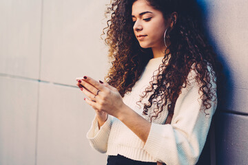 Obraz premium Cropped image of young female person with beautiful curly hair watching interesting video in website on telephone outdoors.Pondering hipster girl checking account balance on telephone outside