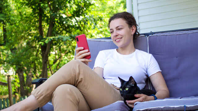 Smiling young woman relax sit on sofa with black pet cat on home terrace,using phone texting chat with friend.Happy brunette girl on couch hold smartphone shop online,check application,social media