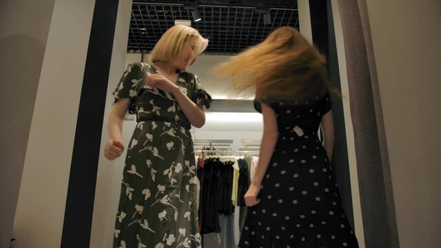 Two girls tried on new dresses in fitting and spinning and show off their choice in front of each other. Modern fitting rooms in the clothing and footwear store in mall.