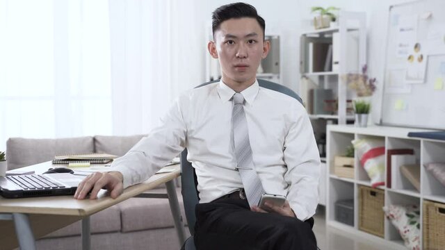 front view male employee is sitting with smartphone in hand, lost in thought. guy wearing shirt and tie facing camera is tapping on desk with finger , looking absent-minded