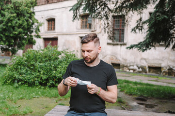 Obraz premium A bearded man holds a face mask in his hands. A man on the street is thinking whether to wear a medical disposable mask. People do not want to wear masks.