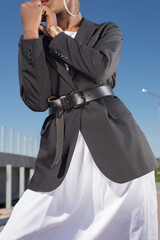 close-up details of a female business black jacket, on a waist a leather belt on an African fashion model, against a blue sky. Style, design of women's clothing. Black fashion girl
