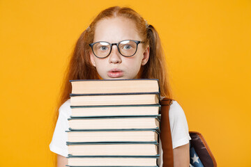 Young fun redhead girl 12-13 years old in white t-shirt eyeglasses backpack hold big stack school textbook notebook book isolated on yellow background children studio portrait Kids education concept