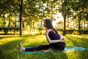 a brunette girl performs the Marichiasana pose in nature at sunset