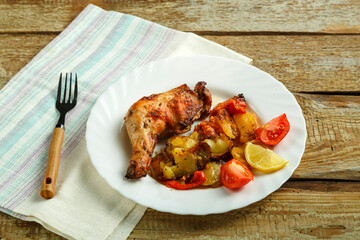Baked chicken leg with potatoes and tomato on a white plate with a fork on a wooden table.