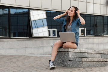 Young fashionable girl uses laptop outdoors and listens to music