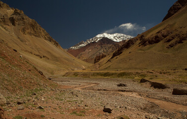 Fototapeta na wymiar Seven summits. View of a stream flowing across the rocky and arid valley with mountain Aconcagua snowy peak in the background.