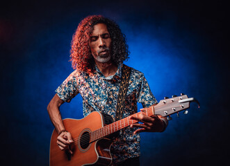 Talented hispanic musician in a hawaiian shirt playing guitar on a dark illuminated by blue and red light. Concept of music, hobby, festival