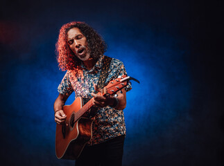Hispanic musician emotionally singing and playing guitar on a dark lluminated by blue and red light. Concept of music, hobby, festival