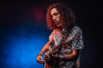 Middle aged hispanic man musician in a hawaiian shirt playing guitar on a dark illuminated by blue and red light. Concept of music, hobby, festival