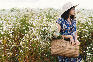 Obraz premium Stylish young woman in blue vintage dress and hat gathering white wildflowers in straw basket in meadow. Beautiful girl holding white daisies bouquet. Tranquil summer in countryside