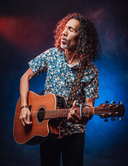 Fototapeta premium Talented hispanic musician in a hawaiian shirt playing guitar on a dark illuminated by blue and red light. Concept of music, hobby, festival