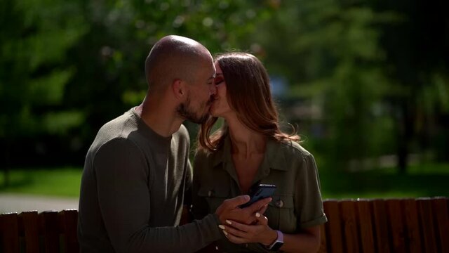 loving pair is kissing and viewing pictures in smartphone, sitting in park
