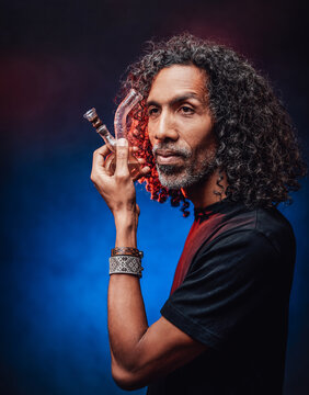 Middle aged hispanic male with long curly hair smoking from bong on a dark background illuminated by blue light