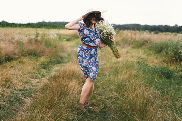 Beautiful girl having fun and laughing in windy field with big daisies bouquet. Authentic summer in countryside. Young woman in blue vintage dress and hat dancing with white wildflowers in meadow.