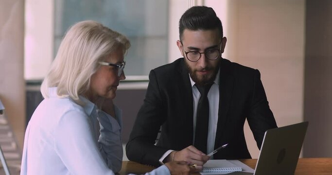 Young arabic eastern ethnic male mentor trainer manager wear suit consult mature old female client teach middle aged intern new worker explain deal benefit computer work on laptop at business meeting