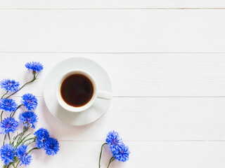 White cup of coffee and blue cornflowers on a vintage white wooden background. Lovely summer flat lay. Copy space for your text and product.