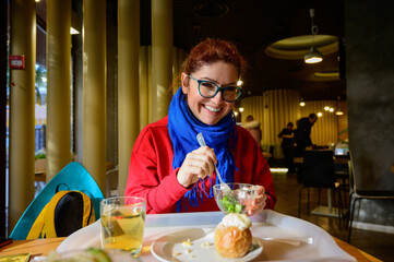 Red-haired caucasian woman eating salad in a cafe. Pensive romantic girl in glasses and a blue scarf is having lunch.