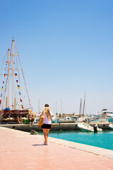 Young blonde woman is walking along port with parked boats.