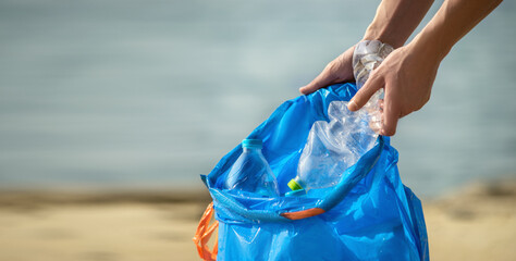Hand picking up plastic bottles into blue trash bag. Cleaning on the beach. Copy space