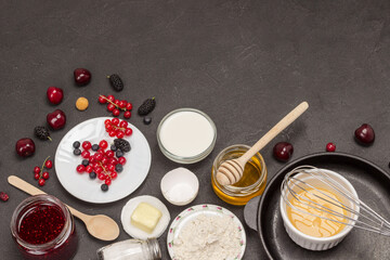 Bowl with beaten eggs, metal whisk in frying pan. Berries, flour in plate. Berries, flour in plate. Black background.