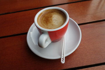 Cup of coffee with plastic spoon on wooden table. Tasty foam, sweet red white ceramic cup top view
