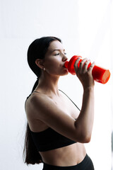 Vertical photo of a female athlete who drinks protein or water from a red shaker on a white...