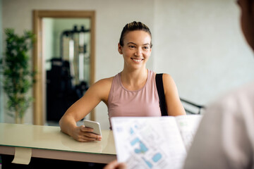 Naklejka premium Happy sportswoman using smart phone while talking to receptionist at health club.