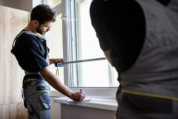 Obraz premium Our service is the best. Two professional workers in uniform using tape measure while measuring window and making notes for installing blinds indoors. Construction and maintenance concept