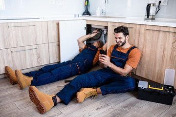 Young repairman, worker in uniform sitting on the floor, using smartphone while his experienced colleague fixing sink pipe indoors. Repair service, new generation concept