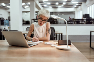 Hard working day. Young, busy tattooed blonde woman in eyewear making some notes and looking at camera while sitting alone in the modern office