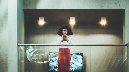 A serious young African-American businesswoman in a white shirt and red skirt is reading an email on her smartphone while leaning on chrome railing of a balcony in an office open-space area