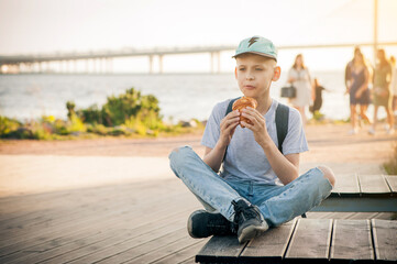 The boy sits on a bench on the waterfront with his legs crossed and eats a croissant. A teenager in a blue cap and T-shirt eats pastries on the street and looks thoughtfully into the distance.