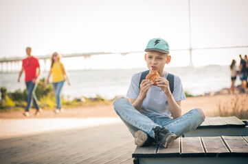 The boy sits on a bench on the waterfront with his legs crossed and eats a croissant. A teenager in the street eats pastries and looks thoughtfully into the distance. People are passing by.