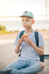 The boy sits on a bench on the waterfront and eats a croissant. A teenager in cap and T-shirt eats pastries on the street and looks thoughtfully into the distance. Vertical photo